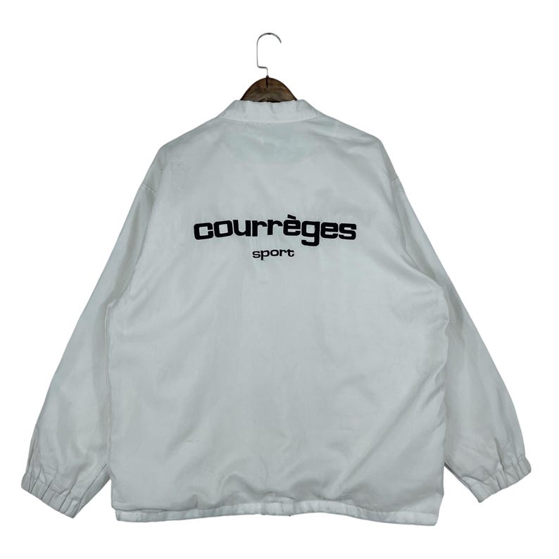 May include: White jacket with the text "courr&egrave;ges sport" in black on the back. The jacket has a collar and long sleeves. The jacket is hanging on a wooden hanger.