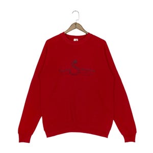 Op de afbeelding: Een felrood sweatshirt met lange mouwen en een ronde hals, voorzien van een donker geborduurd motief op de borst. Het ontwerp toont een gestileerde letter S in de vorm van een slang, vergezeld van de tekst "Sailing Souvenirs" in cursief.