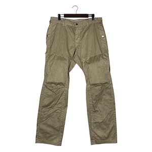 Può includere: Pantaloni color kaki appesi a una gruccia nera. I pantaloni presentano un design unico con toppe rinforzate sulle ginocchia e chiusura con bottoni. Il tessuto sembra essere un materiale leggero, adatto all'abbigliamento casual.