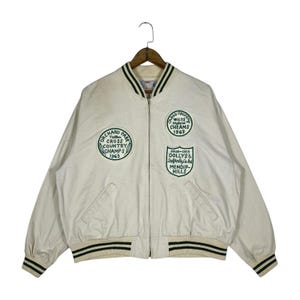 Pode incluir: Casaco vintage creme com acabamento listrado verde e preto. Apresenta patches bordados com texto, incluindo "Orchard Park Cross Country Champs 1963" e "Wilts Cheams 1963".