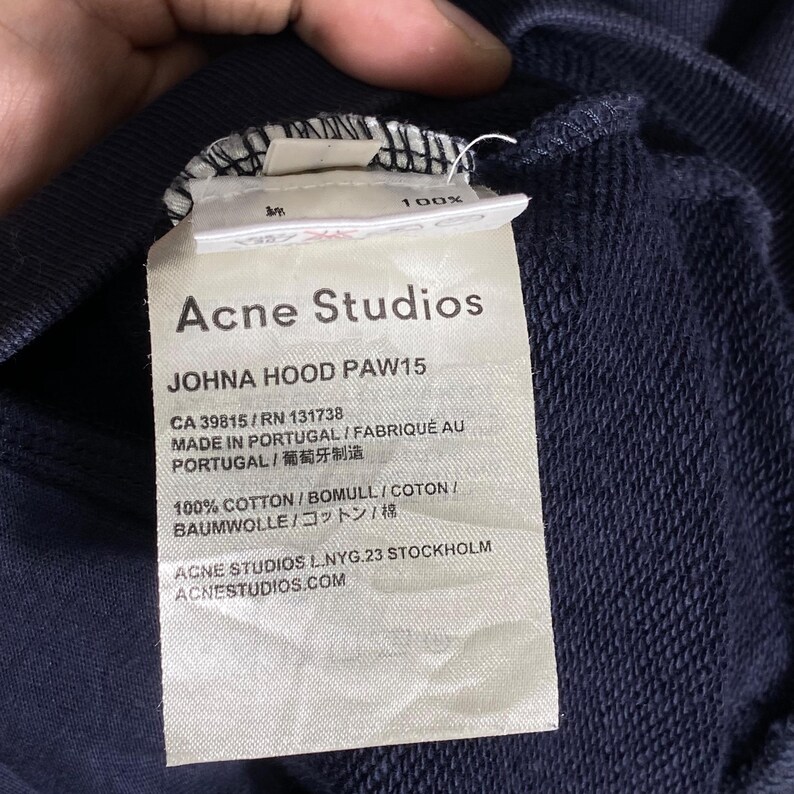 May include: Close-up of a navy blue garment tag. The tag reads "Acne Studios" and "JOHNA HOOD PAW15". Additional text includes "MADE IN PORTUGAL" and "100% COTTON". The tag is white with black text.