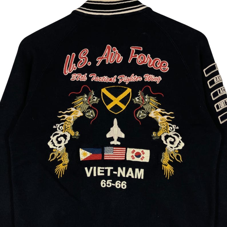 May include: Black bomber jacket with embroidered "U.S. Air Force" text, dragons, flags, and "VIET-NAM 65-66" text. The jacket has a white and black striped collar and cuffs. The back of the jacket features a variety of patches.