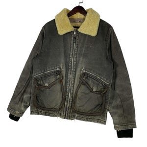 May include: A grey denim jacket with a cream shearling collar. The jacket has a zip-up front, two front pockets with flaps, and black ribbed cuffs. It is suspended from a wooden hanger.