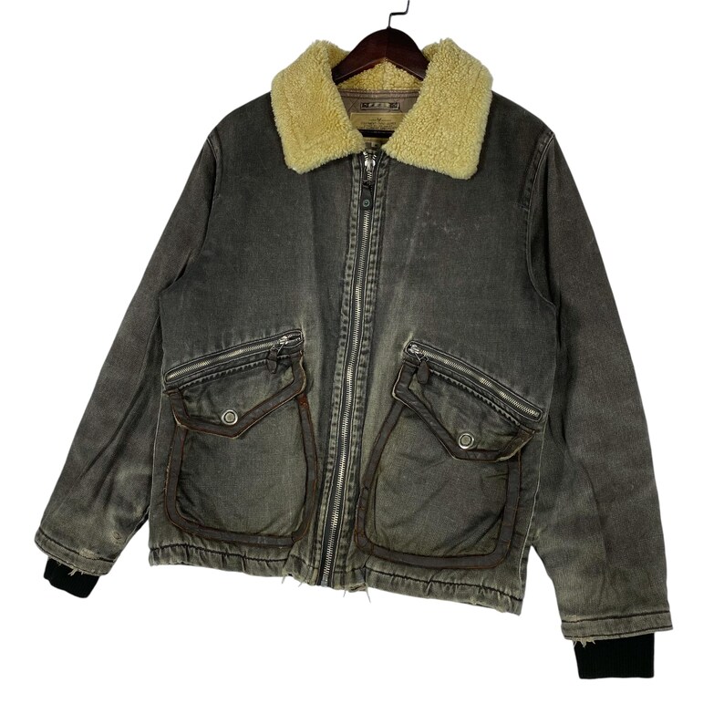 May include: A grey denim jacket with a cream shearling collar. The jacket has a zip-up front, two front pockets with flaps, and black ribbed cuffs. It is suspended from a wooden hanger.