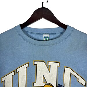 May include: A light blue crewneck sweatshirt featuring the letters UNC in large white block font with a thin yellow outline. The garment has a ribbed collar and a small rectangular brand tag inside the neckline with a green and white graphic.