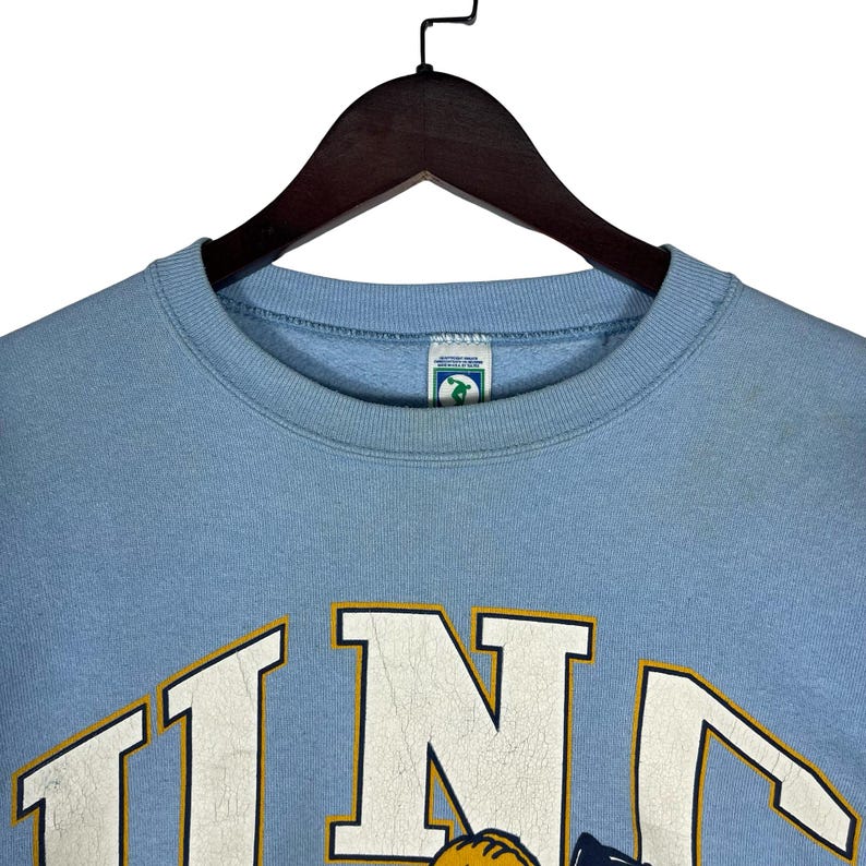 May include: A light blue crewneck sweatshirt featuring the letters UNC in large white block font with a thin yellow outline. The garment has a ribbed collar and a small rectangular brand tag inside the neckline with a green and white graphic.