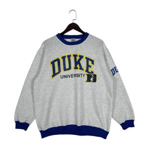Può includere: Una felpa grigio melange con la scritta "DUKE" in lettere blu e gialle sul petto. La parola "UNIVERSITY" è sotto. La felpa ha finiture blu sul colletto, sui polsini e sull'orlo. Il logo Duke è sulla manica.