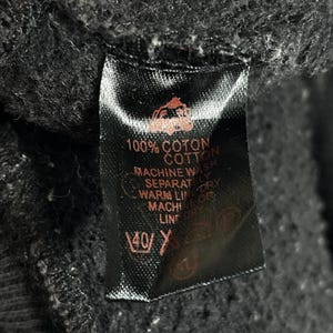 May include: A black clothing tag with white text indicating the garment is 100% cotton and machine washable. The tag also includes washing instructions, such as washing separately and line drying.