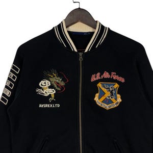 May include: Black bomber jacket with a white and cream striped collar and cuffs. Features embroidered dragon and U.S. Air Force patches. The jacket has a zipper closure and the text "AVIREX LTD".