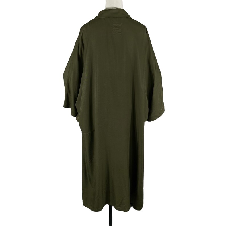 Puede incluir: Chaqueta larga verde oliva estilo kimono con mangas anchas y cuello. Vista trasera de la prenda sobre un maniqu&iacute; blanco. La tela parece ser un material ligero.