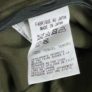 Puede incluir: Primer plano de una etiqueta blanca con texto negro y s&iacute;mbolos de cuidado. La etiqueta dice "FABRIQUE AU JAPON MADE IN JAPAN" y "100% TENCEL TENCEL". Tambi&eacute;n se incluyen instrucciones de lavado. La etiqueta est&aacute; adherida a una tela verde oliva.