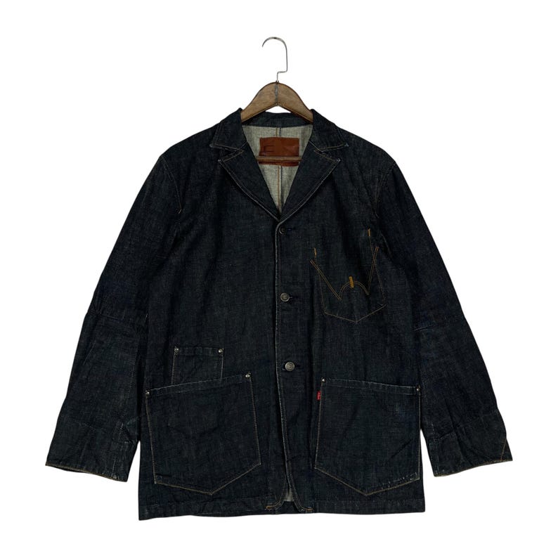 Puede incluir: Chaqueta vaquera azul oscuro con dise&ntilde;o de blazer. Cuenta con solapa con muescas, cierre de tres botones y m&uacute;ltiples bolsillos, incluidos dos grandes bolsillos de parche en la cintura. Una peque&ntilde;a etiqueta roja es visible en un bolsillo. La chaqueta est&aacute; colgada de una percha de madera.