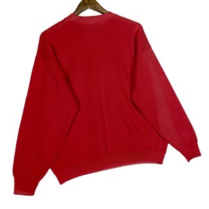 May include: A solid red sweatshirt hangs from a wooden hanger. The sweatshirt has long sleeves and a crew neck. The fabric appears to be a medium weight, suitable for cooler weather. The garment is clean and appears to be in good condition.