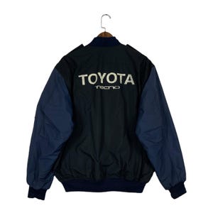 May include: A navy blue and black bomber jacket featuring the word TOYOTA in large white letters across the upper back, with the word TECNO printed in smaller white letters directly underneath. The jacket has a dark blue collar and cuffs.