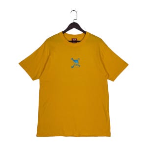 May include: A mustard yellow short-sleeve t-shirt featuring a small, centered blue graphic of a stylized creature. The shirt has a crew neckline and is displayed on a black plastic hanger against a plain white background.