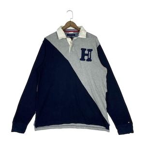 May include: Long-sleeved rugby shirt in navy blue and gray color block design. Features a white collar, a navy blue "H" patch, and the Tommy Hilfiger logo. The shirt is hanging on a wooden hanger.