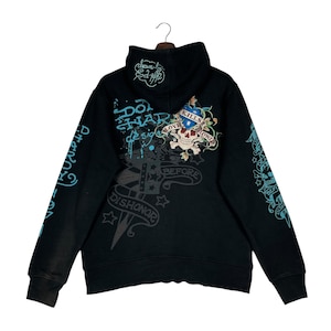 May include: Black hoodie with a skull and floral design on the back, featuring the text "Love Kills Slowly" and "Don Hardy Design". The sleeves have additional text and graphics in a teal color. The hoodie has a drawstring hood.