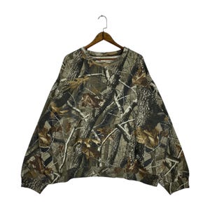 May include: A camouflage-patterned sweatshirt hangs from a wooden hanger. The long-sleeved top features a repeating design of tree branches and leaves in shades of brown, green, and tan. The crew neck sweatshirt is made of a soft material.