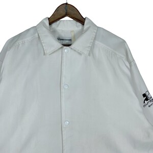 May include: White button-up jacket with a collar, hanging on a wooden hanger. The jacket has a small logo on the sleeve and a label near the collar. The jacket is made of a lightweight material.