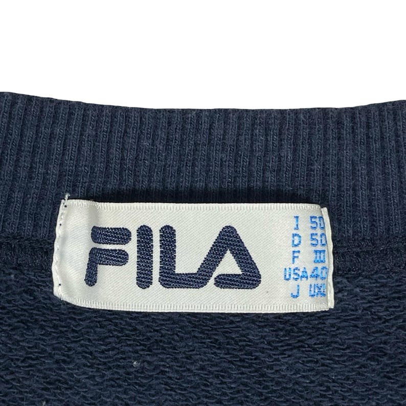 May include: A dark navy blue garment featuring a white interior neck label with the navy blue FILA logo printed on the left side. The label includes size information for various regions, including USA 40, printed in blue text on the right side.
