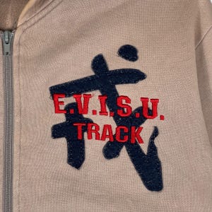 Puede incluir: Chaqueta deportiva beige con cremallera y un gran dise&ntilde;o bordado en azul marino y rojo en la parte delantera. El dise&ntilde;o incluye el texto "E.V.I.S.U. TRACK" en letras rojas.