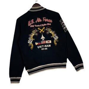 May include: Black bomber jacket with white and beige striped trim. The back features embroidered text "U.S. Air Force" and "VIET-NAM 65-66" with a fighter jet, flags, and dragon designs. The jacket has a ribbed collar and cuffs.