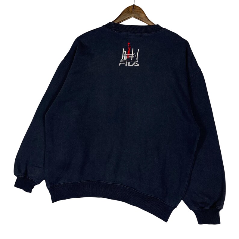 Puede incluir: Sudadera azul marino con cuello redondo y mangas largas. La parte trasera presenta un dise&ntilde;o bordado en blanco y rojo con el nombre de la marca "FILA". La sudadera est&aacute; colgada en una percha de madera.