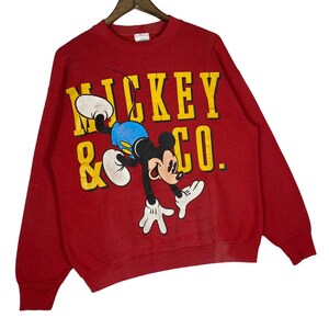 May include: Red sweatshirt featuring a graphic of Mickey Mouse in a playful pose. The text "Mickey & Co." is printed in yellow block letters. The sweatshirt has long sleeves and a crew neck.