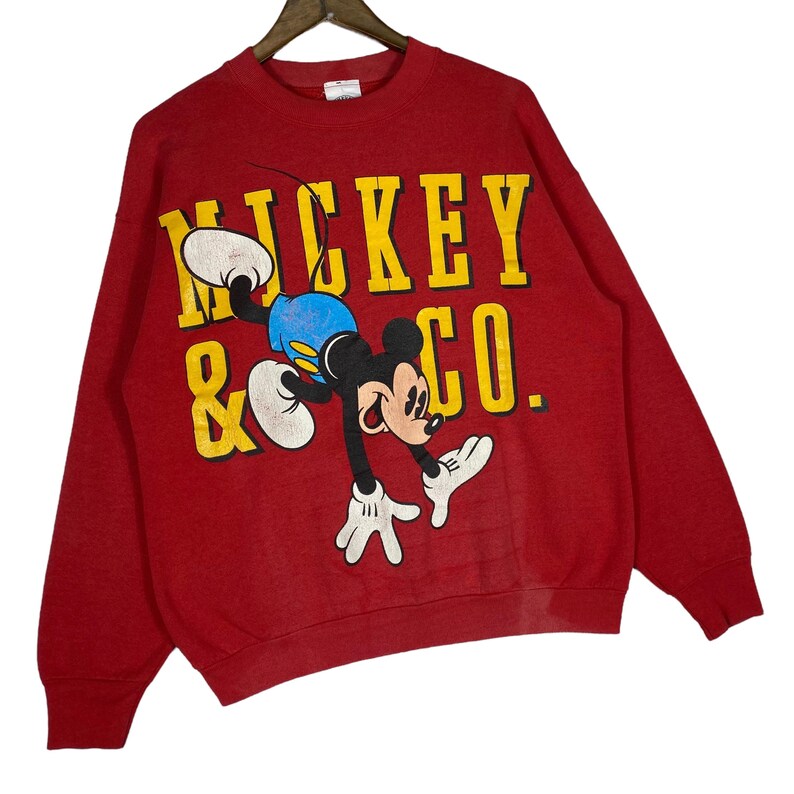 May include: Red sweatshirt featuring a graphic of Mickey Mouse in a playful pose. The text "Mickey & Co." is printed in yellow block letters. The sweatshirt has long sleeves and a crew neck.