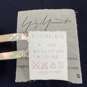 May include: A close-up of a clothing tag with the text "Yohji Yamamoto" in cursive. The tag includes the text "FI-C01-135", "毛 100%", and care symbols. The tag is held by a metal clamp.