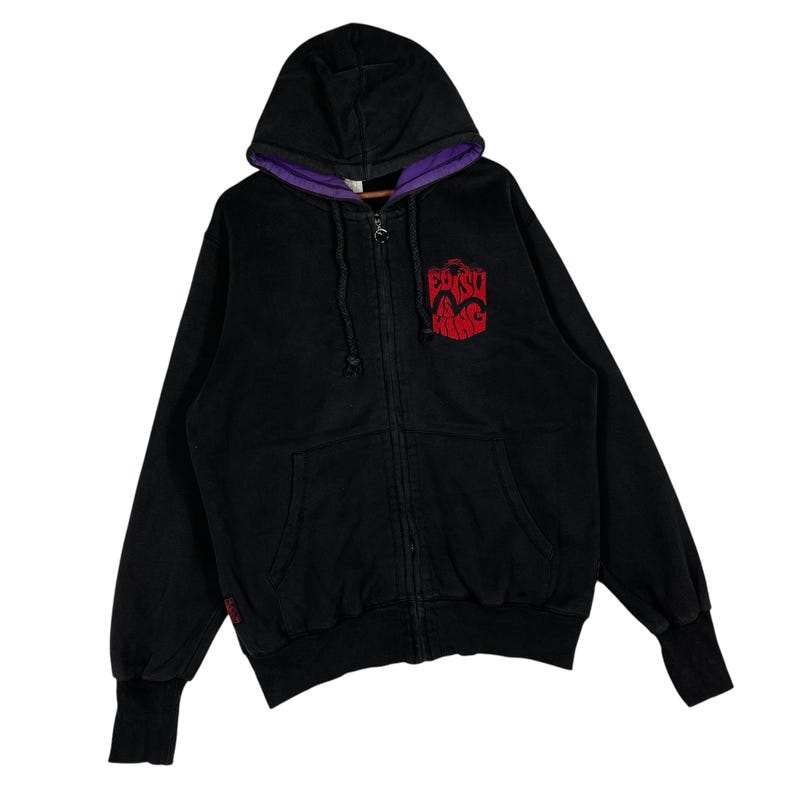 May include: Black zip-up hoodie with a purple-lined hood. The hoodie features a red graphic on the chest. The cuffs and hem are ribbed. The hoodie has a drawstring and a zipper closure.