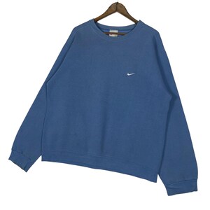 以下が含まれることがあります： 胸に小さな白いナイキのロゴが入った、青いクルーネックのスウェットシャツ。長袖のトップスは木製のハンガーに吊るされています。生地は柔らかく、少し色あせた素材のようです。