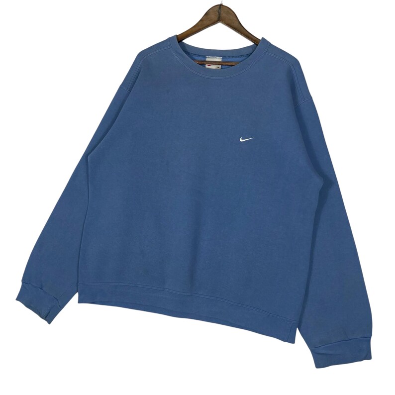 以下が含まれることがあります： 胸に小さな白いナイキのロゴが入った、青いクルーネックのスウェットシャツ。長袖のトップスは木製のハンガーに吊るされています。生地は柔らかく、少し色あせた素材のようです。