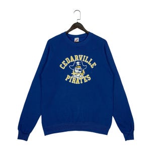 Op de afbeelding: Een koningsblauw sweatshirt met lange mouwen en een afbeelding van een piraat met een ooglapje en een zwaard in de mond. De tekst CEDARVILLE PIRATES is in gele, gebogen letters boven en onder de piratenafbeelding op de borst gedrukt.