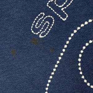 May include: Close-up of navy blue fabric with white embroidered details. The design features a large, stylized letter "S" and a dotted line, possibly part of a larger design. There are dark stains on the fabric.