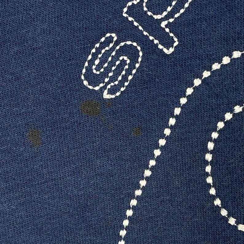 May include: Close-up of navy blue fabric with white embroidered details. The design features a large, stylized letter "S" and a dotted line, possibly part of a larger design. There are dark stains on the fabric.