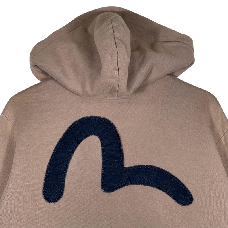 Puede incluir: Una sudadera con capucha de color beige con un logotipo curvo azul oscuro en la espalda. La sudadera con capucha tiene una capucha y mangas largas. El logotipo es un dise&ntilde;o curvo estilizado. La tela parece ser un material suave e informal.
