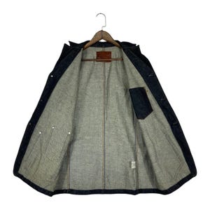 Puede incluir: Chaqueta vaquera azul oscuro con interior gris claro. La chaqueta est&aacute; abierta, revelando el forro interior, los bolsillos y los cierres de botones. Un parche de cuero marr&oacute;n es visible cerca del cuello.