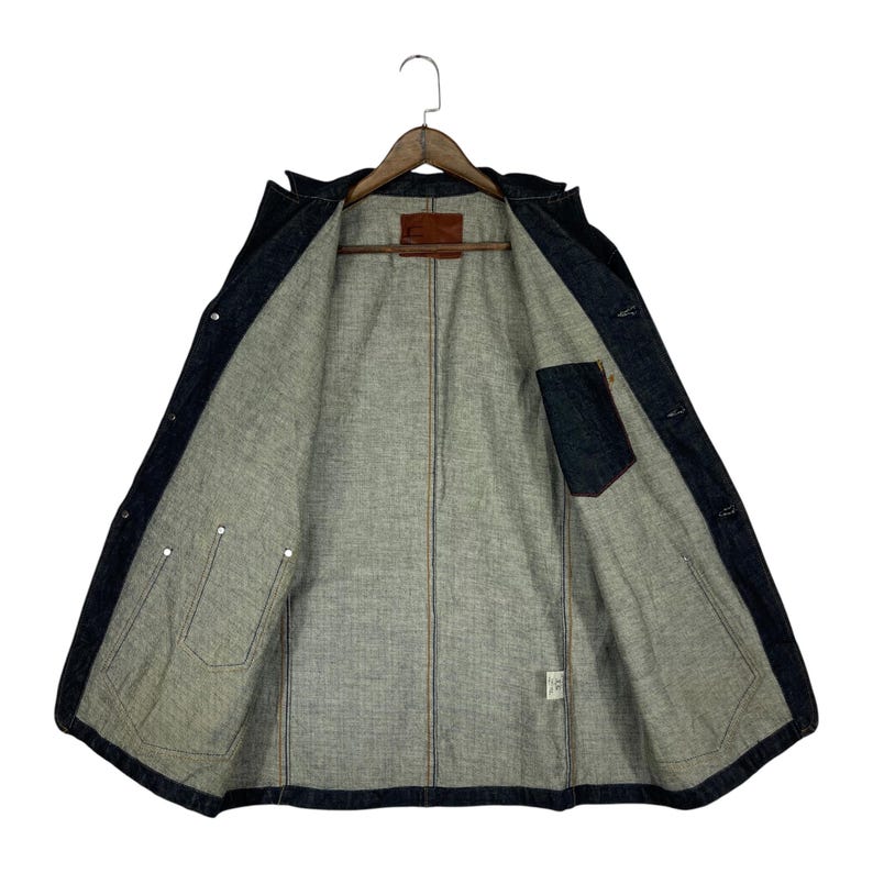 Puede incluir: Chaqueta vaquera azul oscuro con interior gris claro. La chaqueta est&aacute; abierta, revelando el forro interior, los bolsillos y los cierres de botones. Un parche de cuero marr&oacute;n es visible cerca del cuello.
