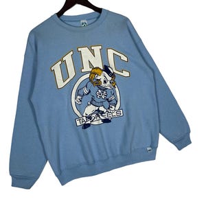 May include: A light blue crewneck sweatshirt featuring the text UNC in large white letters with gold outlines. Below the text is a graphic of a ram mascot wearing a blue sweater with an NC logo, positioned above the text TAR HEELS in a white banner.