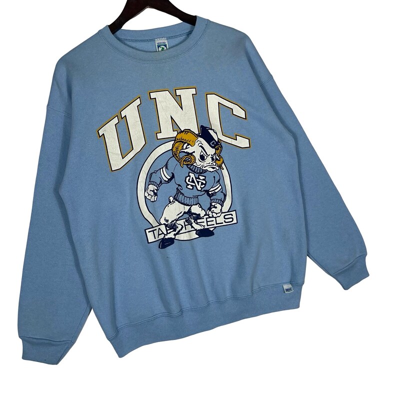 May include: A light blue crewneck sweatshirt featuring the text UNC in large white letters with gold outlines. Below the text is a graphic of a ram mascot wearing a blue sweater with an NC logo, positioned above the text TAR HEELS in a white banner.