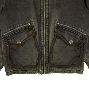 May include: Close-up of a dark grey denim jacket with a full-length zip. The jacket has two large, square pockets with zip closures and a smaller pocket on each side. The pockets are trimmed with a darker brown material, and the jacket has a worn, vintage appearance.