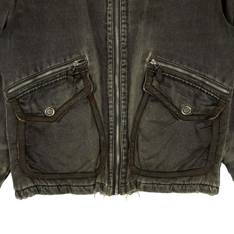 May include: Close-up of a dark grey denim jacket with a full-length zip. The jacket has two large, square pockets with zip closures and a smaller pocket on each side. The pockets are trimmed with a darker brown material, and the jacket has a worn, vintage appearance.