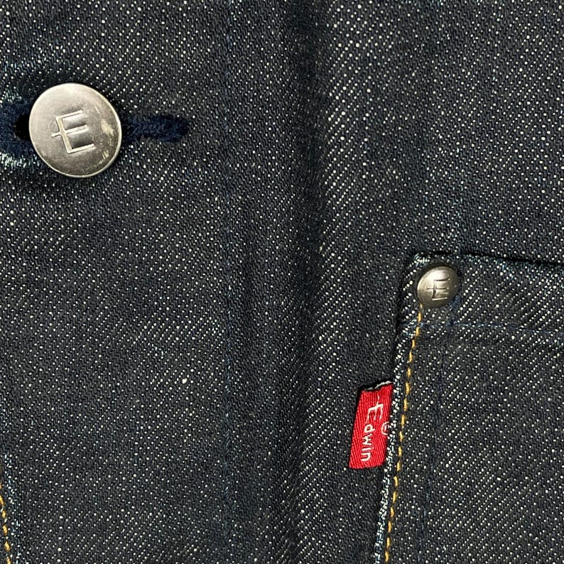 Puede incluir: Primer plano de tejido vaquero azul oscuro con un bot&oacute;n plateado y una peque&ntilde;a etiqueta roja Edwin. El bot&oacute;n tiene un dise&ntilde;o en relieve. El denim tiene un aspecto texturizado.