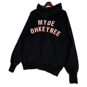 May include: Black hoodie with the text "MYDE OHKEYNEE" in red and white lettering on the back. The hoodie has a hood and long sleeves with ribbed cuffs. The garment is hanging on a wooden hanger.