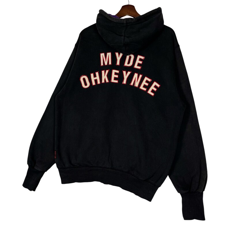 May include: Black hoodie with the text "MYDE OHKEYNEE" in red and white lettering on the back. The hoodie has a hood and long sleeves with ribbed cuffs. The garment is hanging on a wooden hanger.