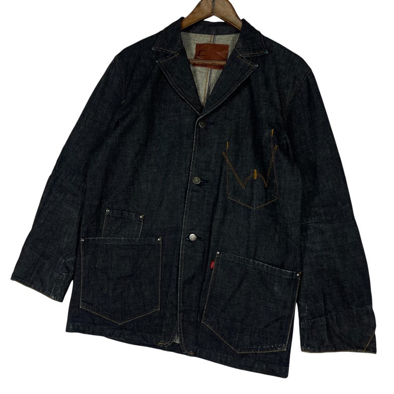 Puede incluir: Chaqueta vaquera azul oscuro con dise&ntilde;o de blazer cl&aacute;sico. Cuenta con tres bolsillos delanteros, un bolsillo en el pecho y cierre de botones. La chaqueta tiene un parche de cuero marr&oacute;n en el interior del cuello y una etiqueta roja en el bolsillo.