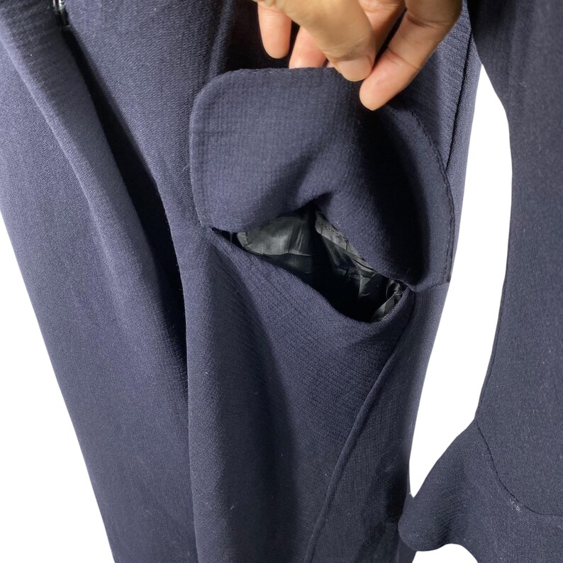 May include: Close-up of a navy blue garment with a pocket. The pocket is open, revealing a black lining. The fabric has a textured appearance. The garment's sleeve is visible on the right side.