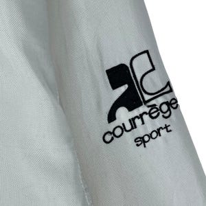May include: Close-up of a white fabric with a black logo that reads "courr&egrave;ge sport". The logo is on the sleeve of a garment. The fabric has a subtle texture.