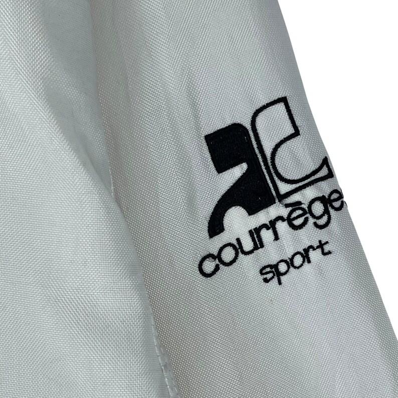May include: Close-up of a white fabric with a black logo that reads "courr&egrave;ge sport". The logo is on the sleeve of a garment. The fabric has a subtle texture.